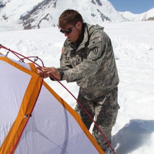 A soldier assembles an orange-and-white expedition dome tent on the snow-covered Kahiltna Glacier below Denali
