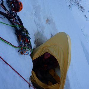 A climber peers out of a yellow single-person bivy shelter dug into a snow ledge on a big alpine wall