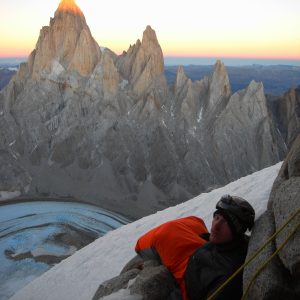 A mountaineer wrapped in an insulated sleeping bag rests during a high-altitude bivouac with Fitz Roy and Cerro Torre lit by sunrise behind him