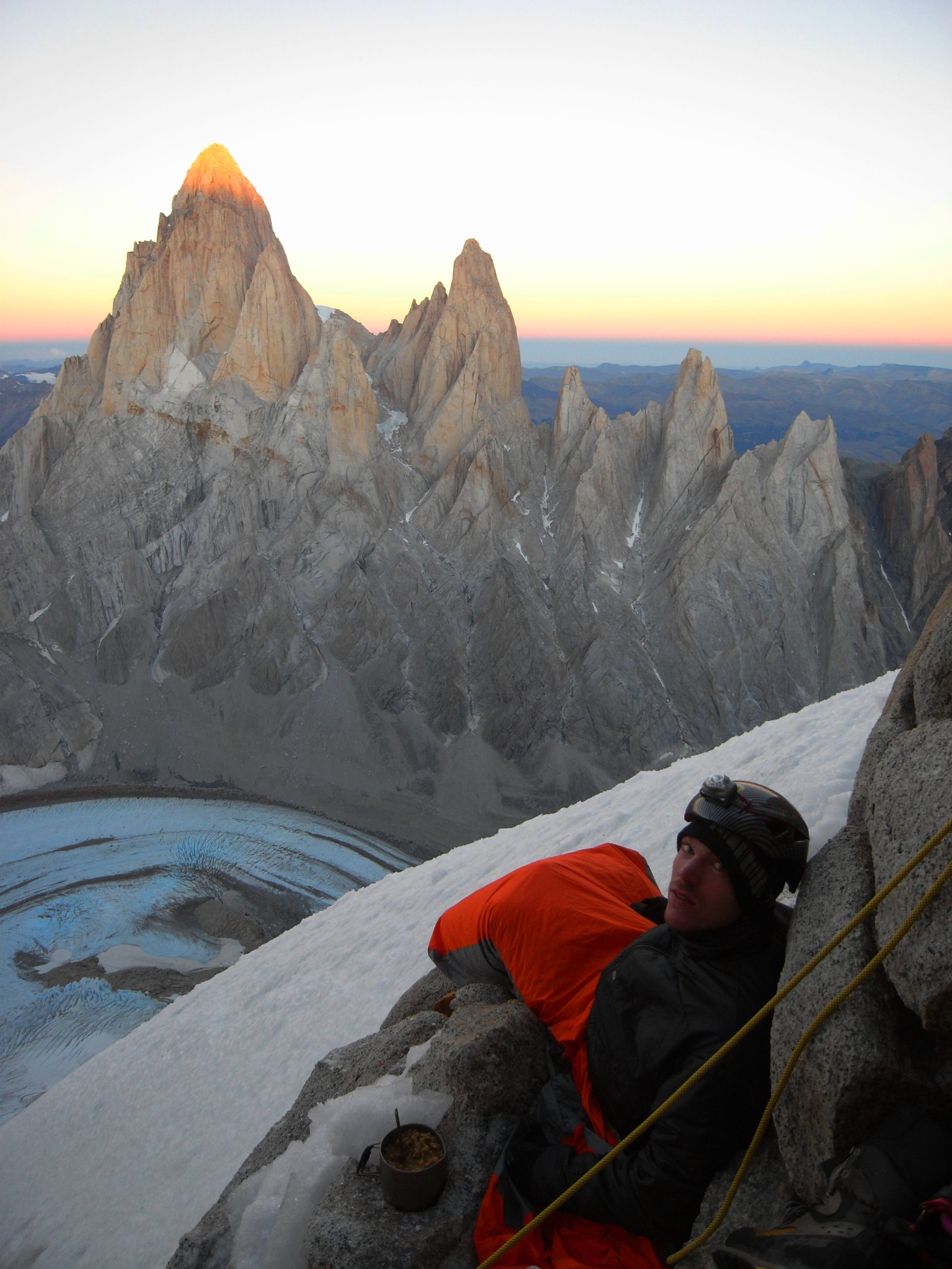 A mountaineer wrapped in an insulated sleeping bag rests during a high-altitude bivouac with Fitz Roy and Cerro Torre lit by sunrise behind him
