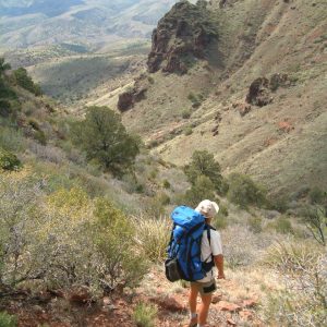 A hiker carrying a large blue internal-frame expedition backpack descends a mountain trail through a desert canyon