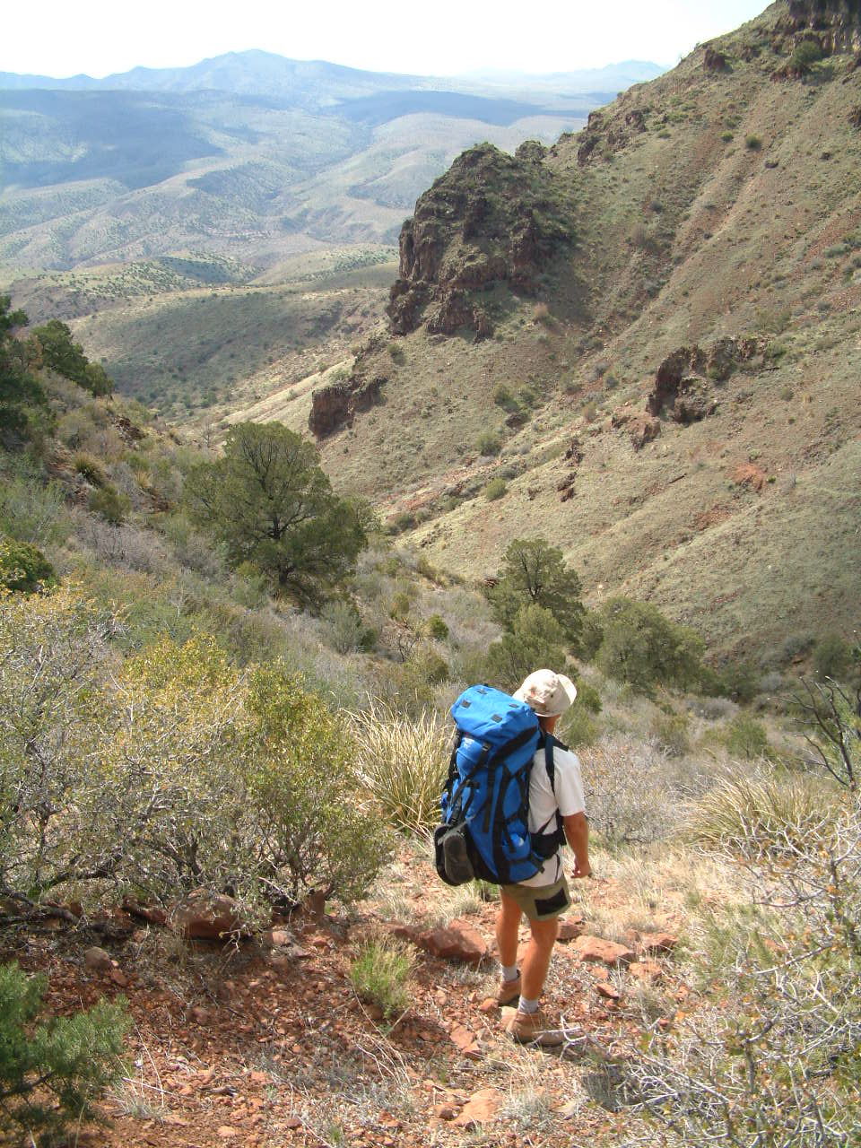 A hiker carrying a large blue internal-frame expedition backpack descends a mountain trail through a desert canyon