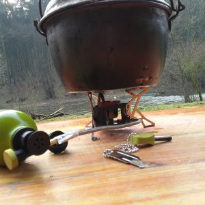 A liquid-fuel camp stove with fuel line connected to an inverted bottle supports a blackened cooking pot at a riverside campsite